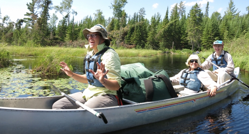 Three people sit in a canoe on calm water. Behind the canoe, lily pads rest on the water, with grass and a line of evergreen trees in the background.